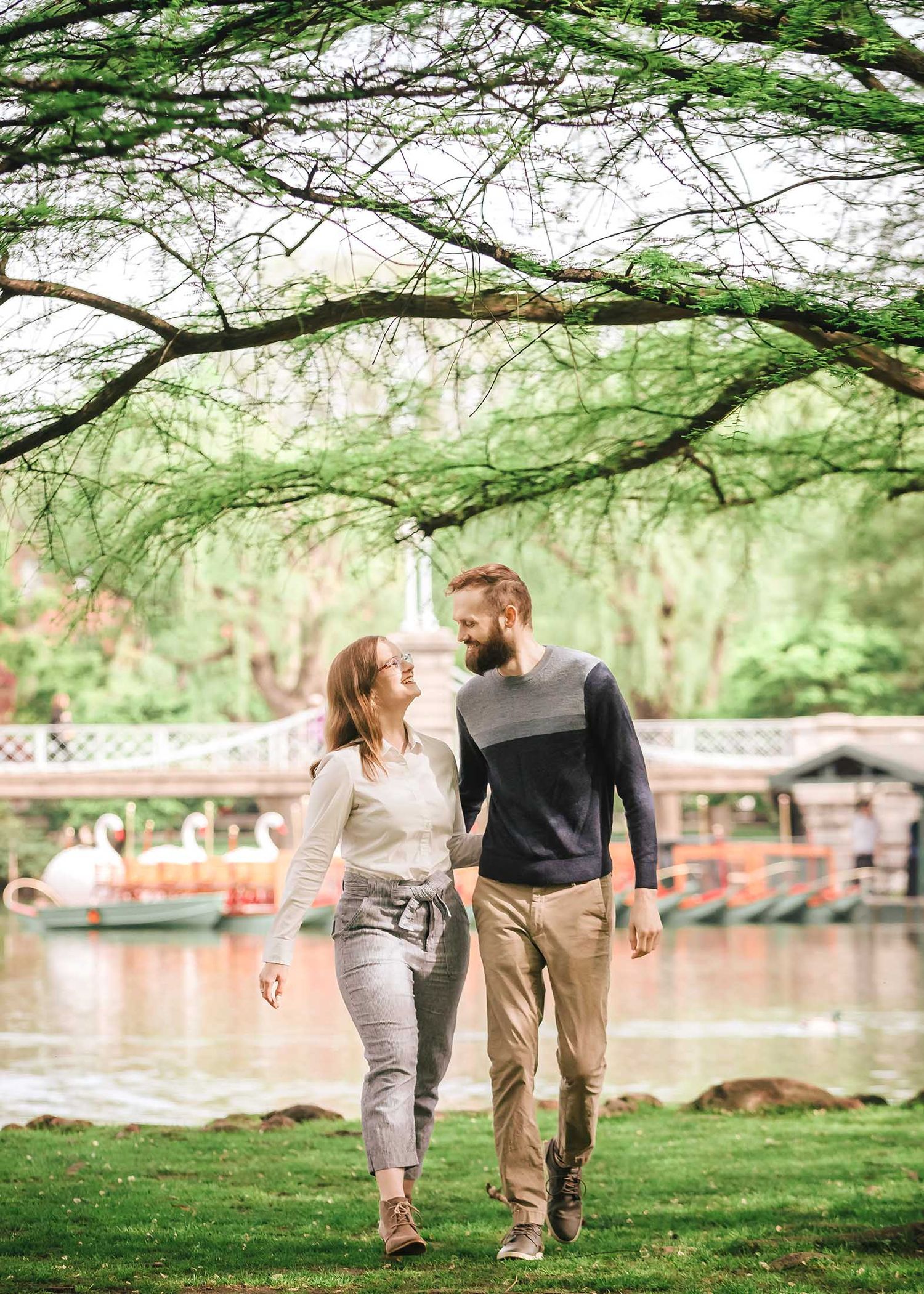Playful candid of couple walking hand in hand beneath greenery near Boston Public Garden Foot Bridge during engagement session
