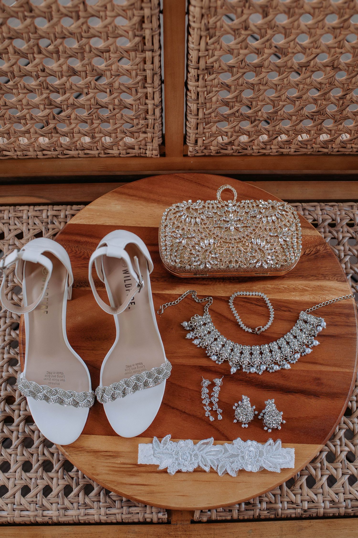 Elegant bridal accessories including white sandals, crystal clutch, jewelry set and garter on wooden circular surface.