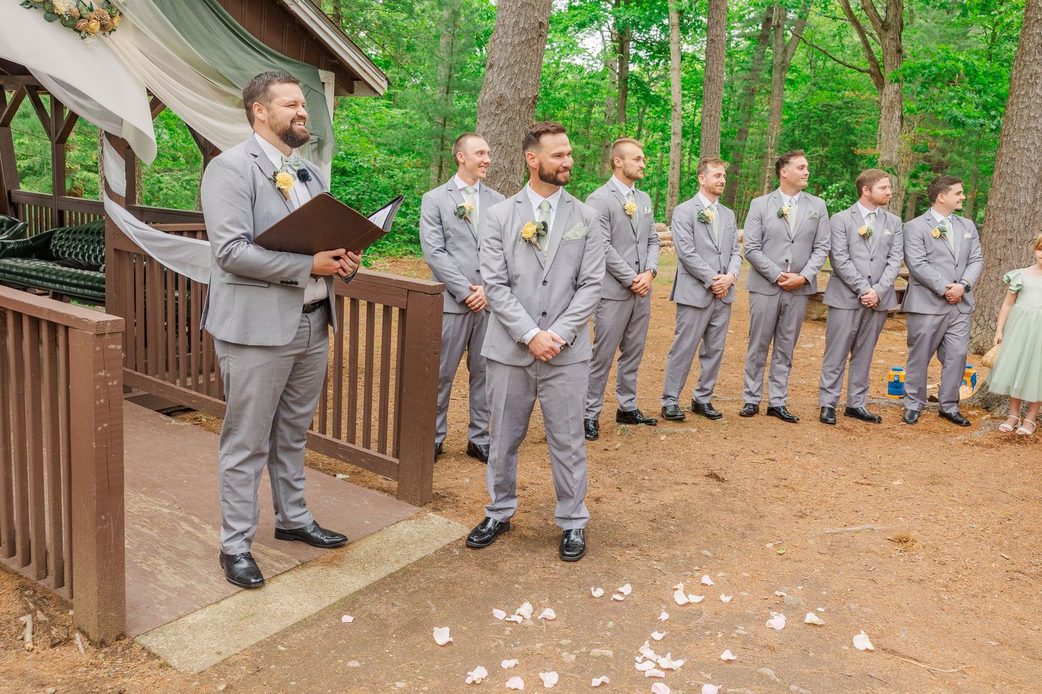 A group wearing matching light gray suits stands on a wooden deck surrounded by trees during a wedding ceremony.