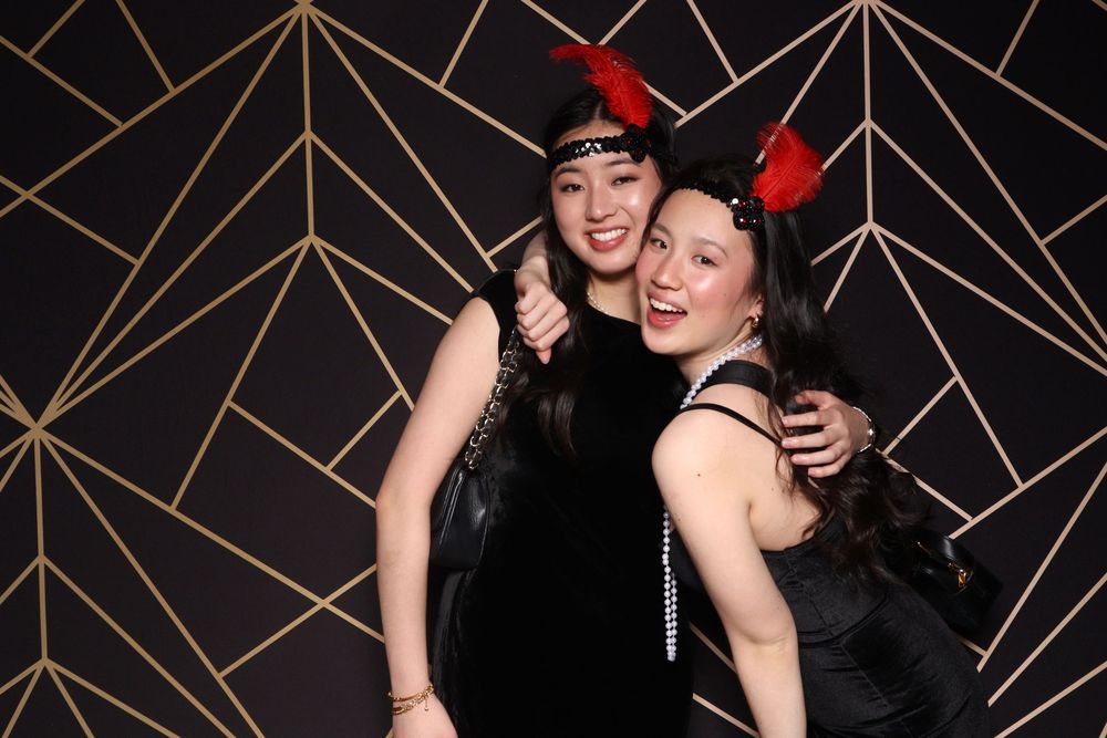 A group poses playfully together in black outfits against a decorative gold geometric pattern wall.