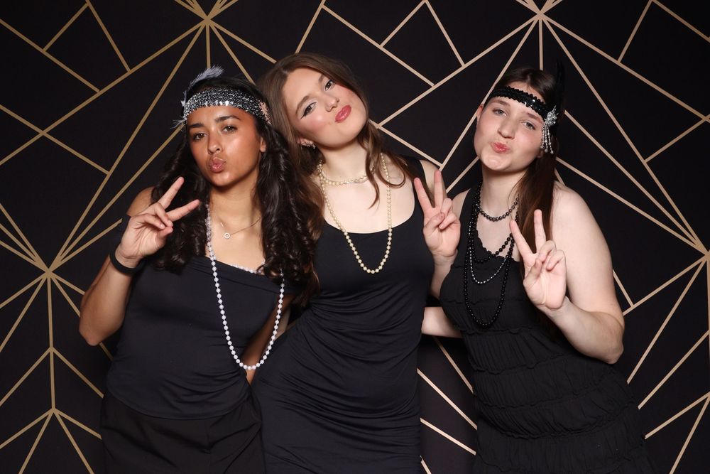 Three friends in black dresses pose against geometric gold backdrop at party.