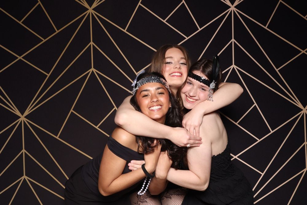 Three friends embrace in a group hug against a black and gold geometric patterned wall.