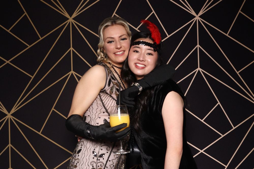 Two partygoers pose together against art deco backdrop at formal event.