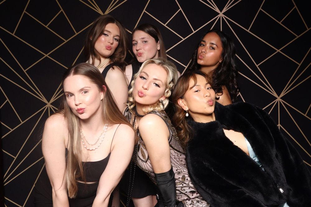 Group of women posing together in dark elegant outfits against a geometric black and gold patterned backdrop.