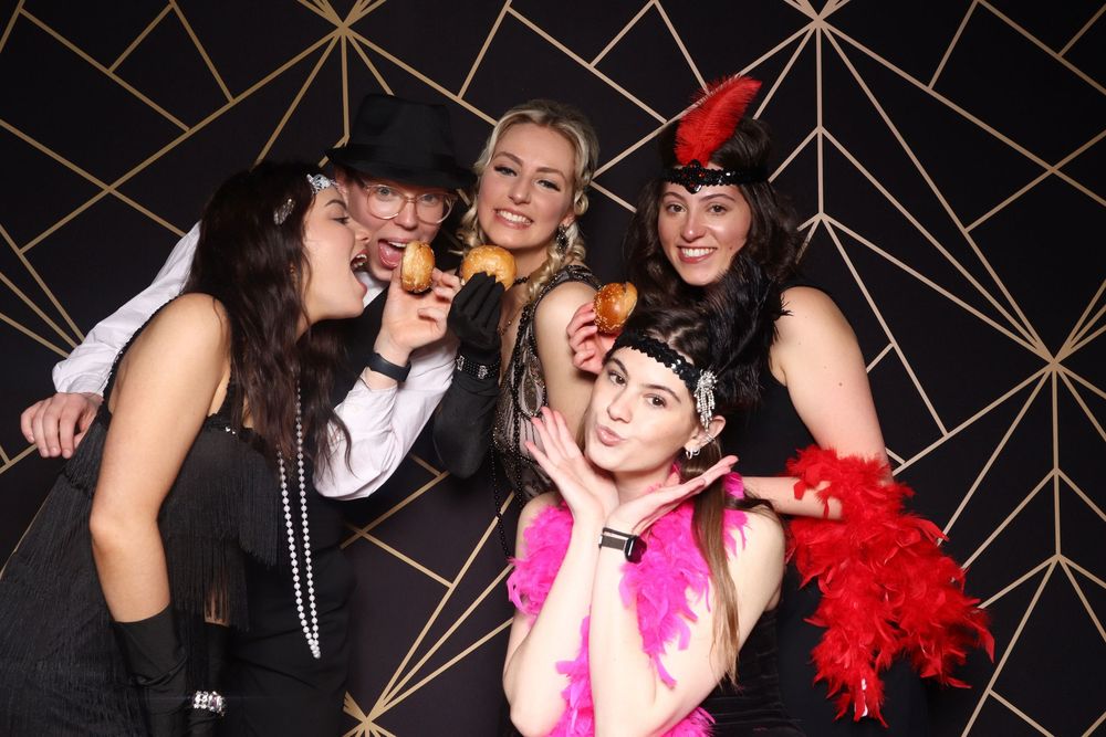 A group poses for fun photos in a party photo booth with props like feather boas against a geometric black and gold backdrop.