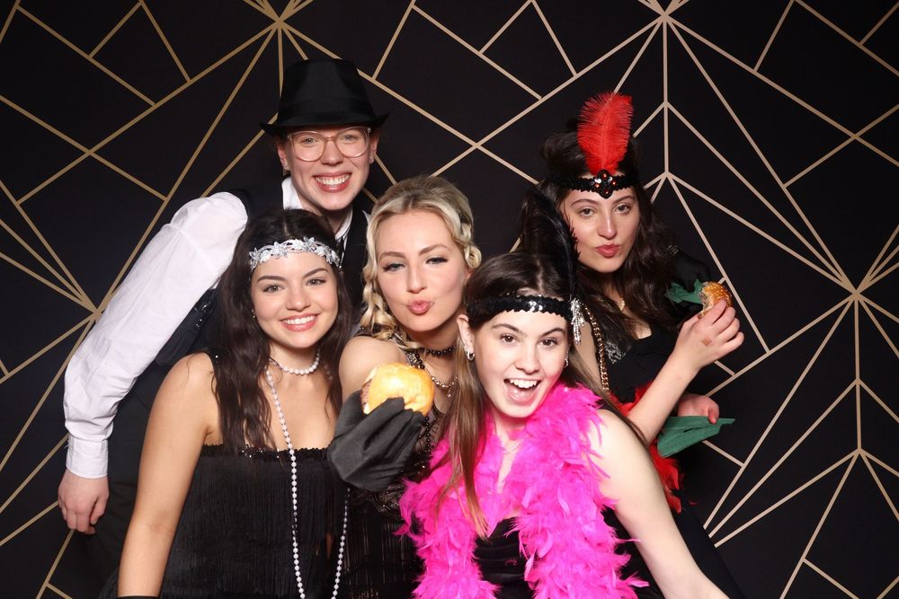 A group poses in a photo booth against a geometric backdrop wearing festive accessories like boas, headbands and hats.