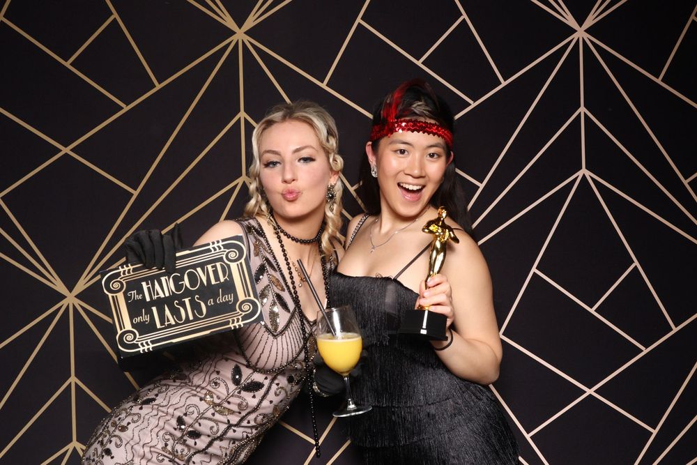 Three party-goers pose together in front of a black and gold geometric wall display at a celebration event.