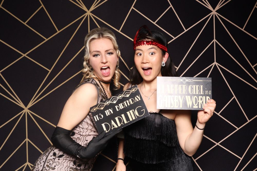 Two friends pose with props against a black and gold geometric backdrop at a photo booth.