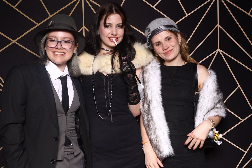 Three people dressed in formal noir style attire pose together against a black and gold geometric backdrop at an event.