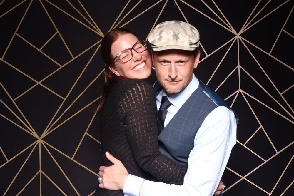 A couple embraces and smiles in front of a geometric black and gold backdrop.