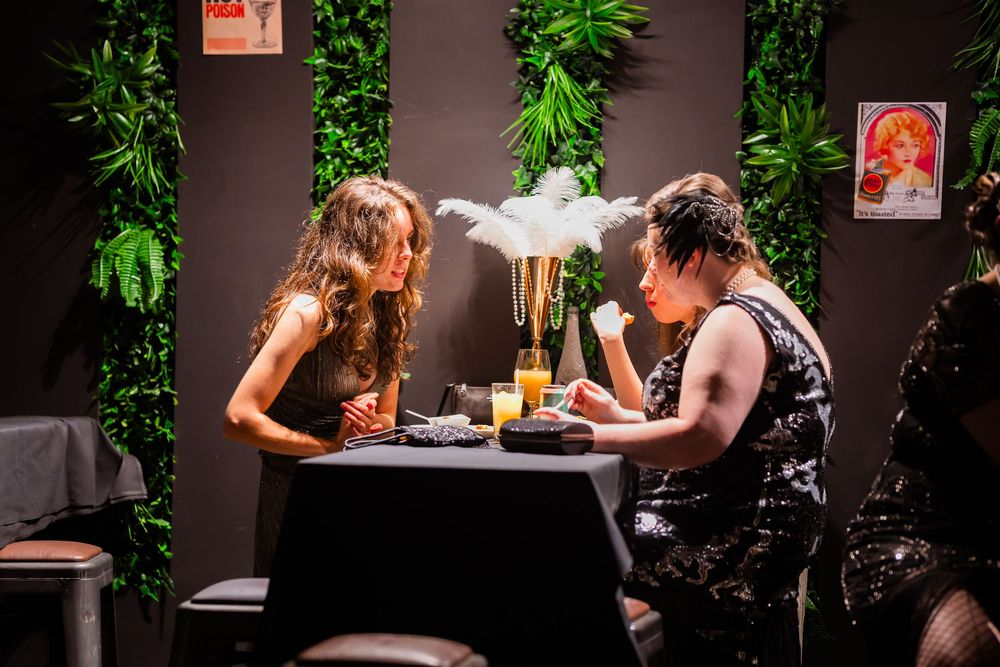 Two people share a meal at an intimate table in a dark restaurant with green plants and framed art on the walls.
