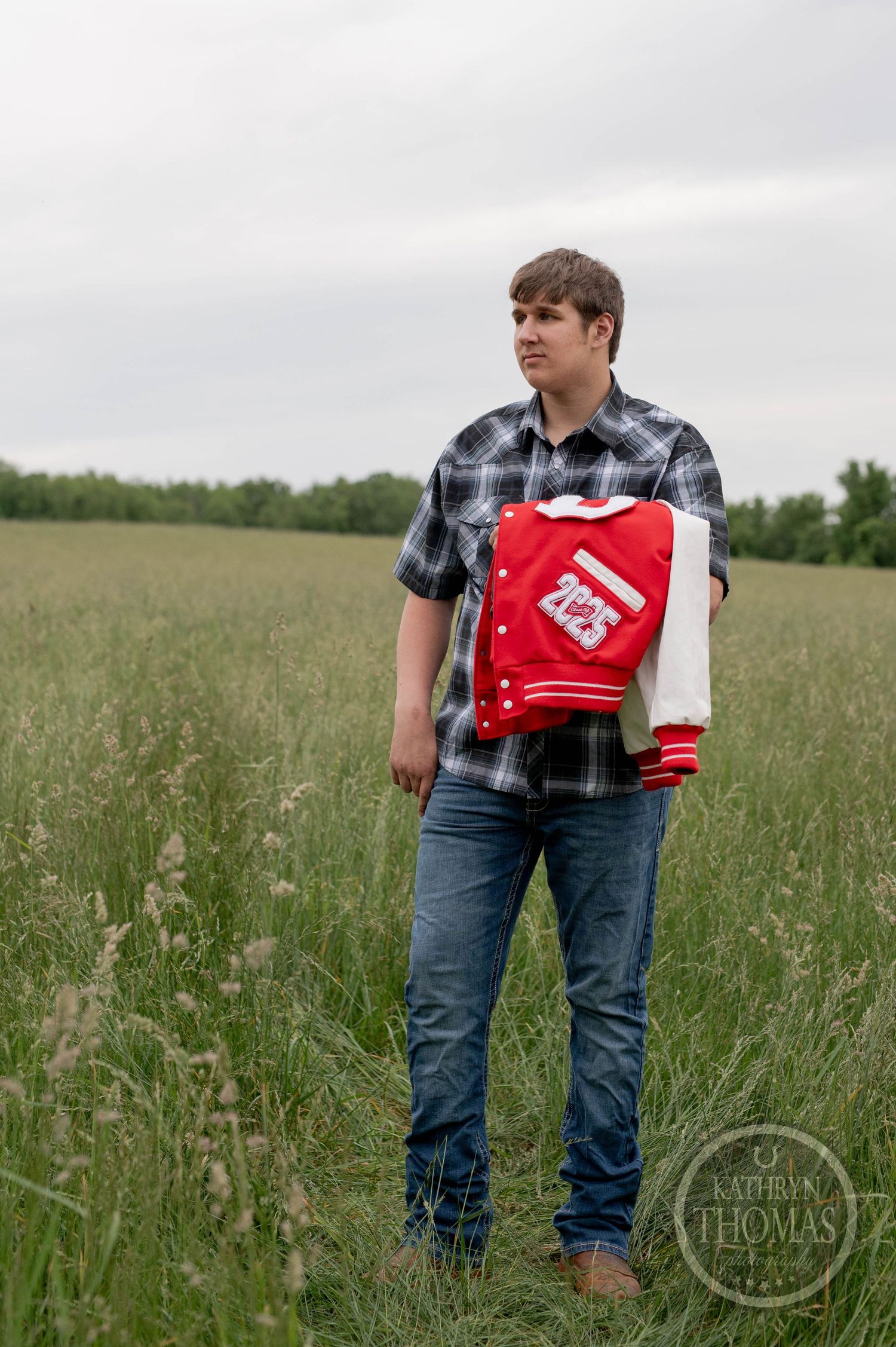 A person in a plaid shirt and jeans stands in a grassy field while holding a red and white varsity jacket.