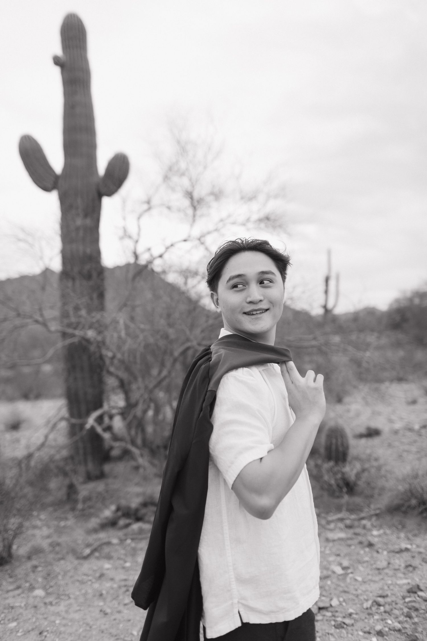 Black and white desert graduate portrait with towering saguaro cactus against cloudy sky.