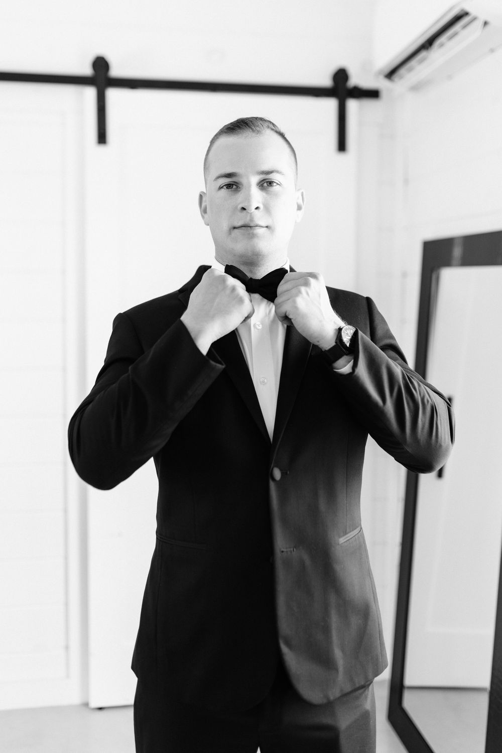 Black and white portrait of groom adjusting his bowtie while standing in front of sliding barn doors.