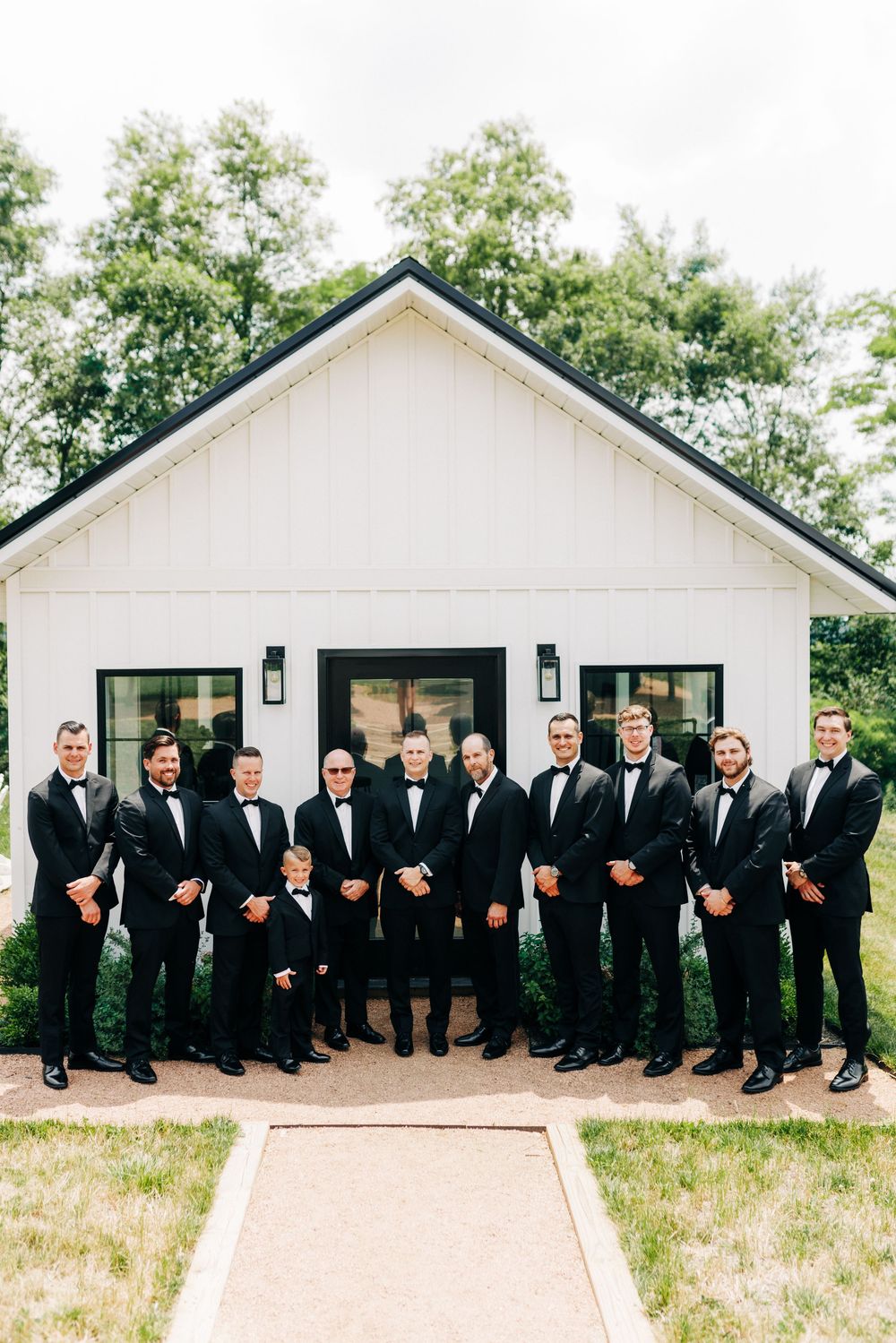 Large group of groomsmen in black suits posing in front of a white chapel building with black trim.