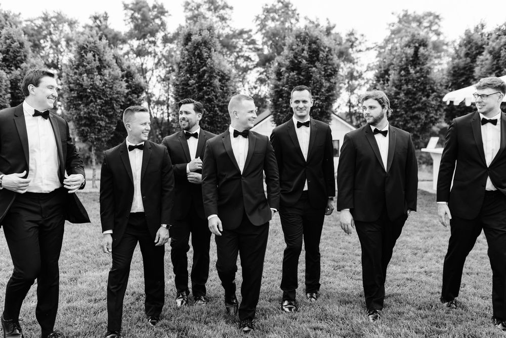 Group of groomsmen in black tuxedos and bowties walking together on grass in elegant wedding party photography.