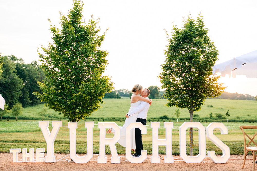 A couple embraces between two green trees with large white letters spelling 'THE YURCHOS' on the ground at sunset.