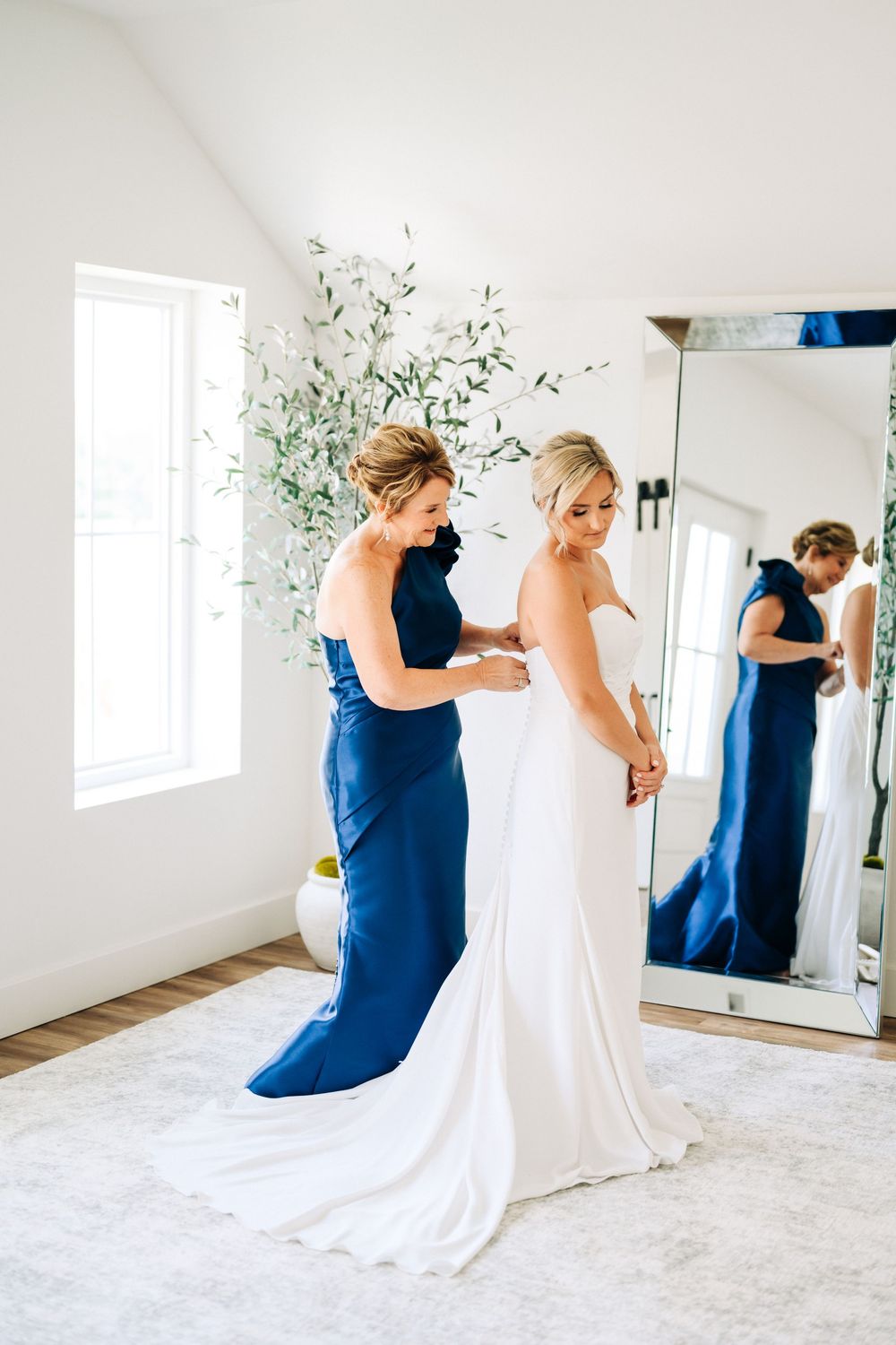 A wedding dress fitting moment showing the elegant white gown being adjusted by an attendant in navy blue.