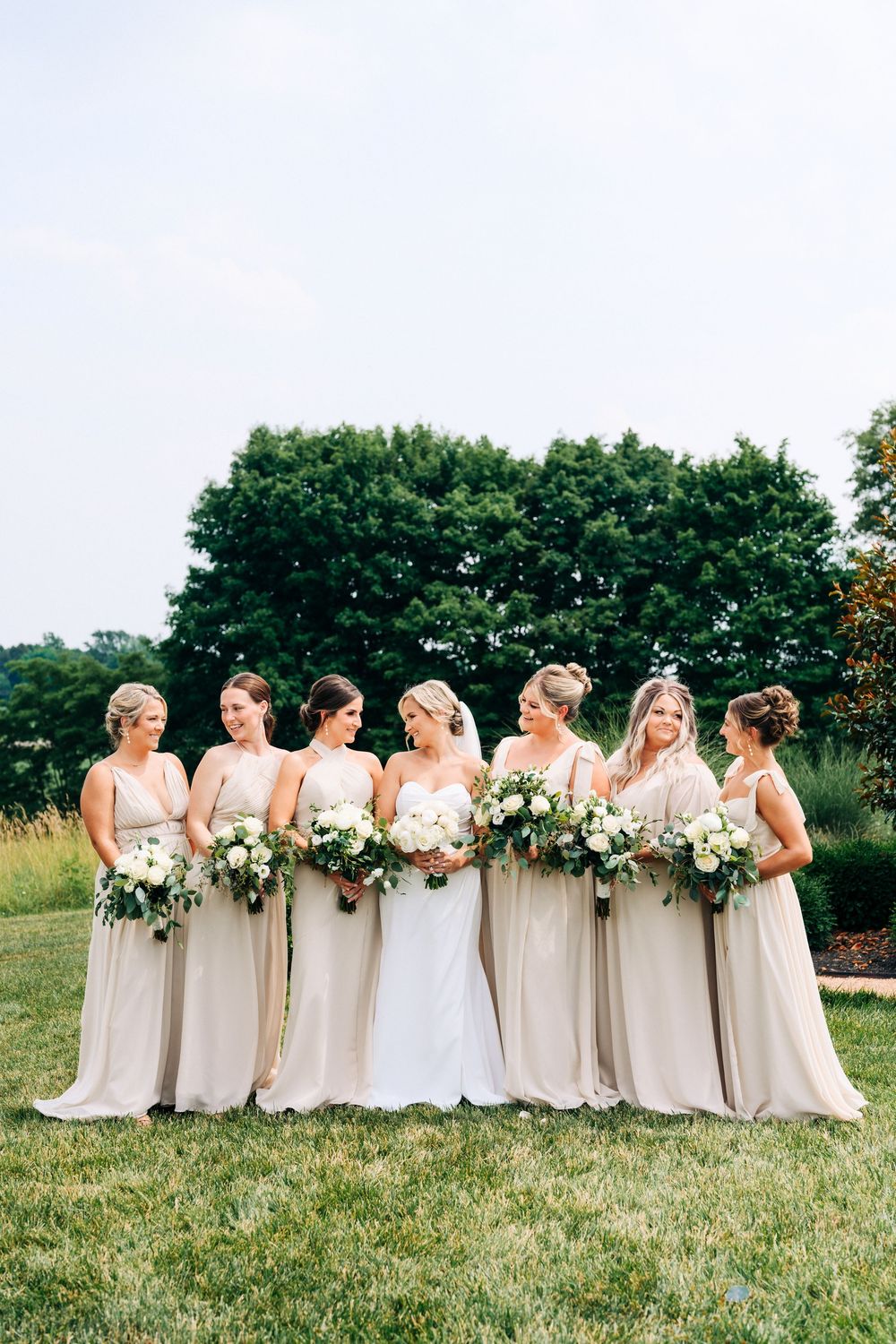 A group of bridesmaids in elegant champagne-colored dresses holding white bouquets pose together on a grassy lawn.