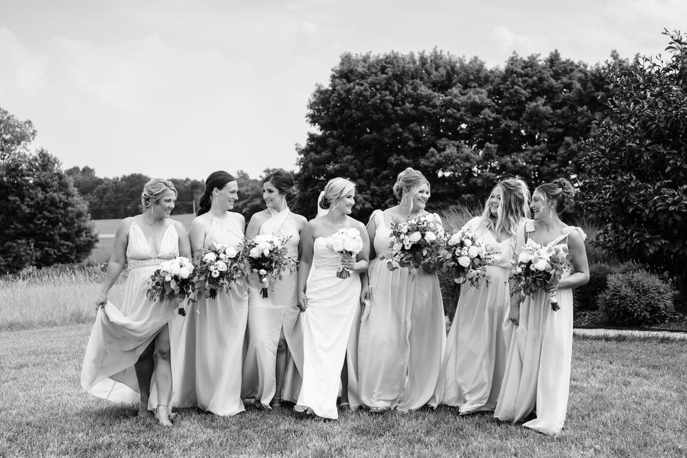 Bridesmaids in flowing light-colored gowns hold bouquets while standing together on grass near trees.