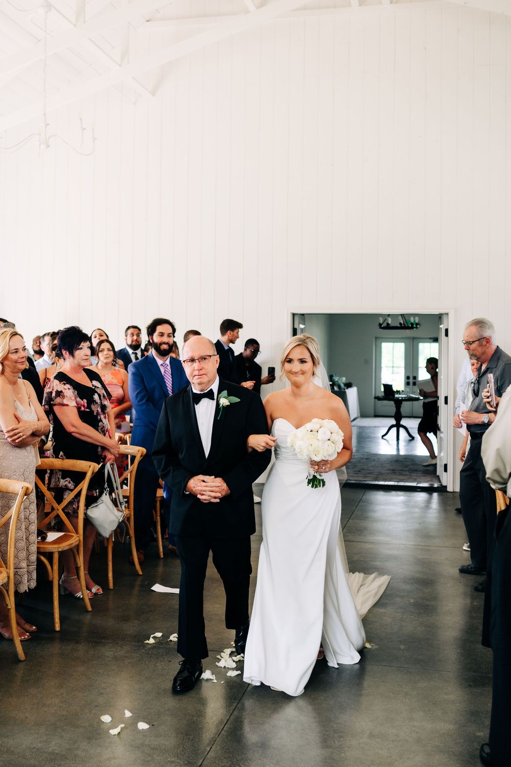 A wedding ceremony processional in a modern venue with guests seated in wooden chairs.