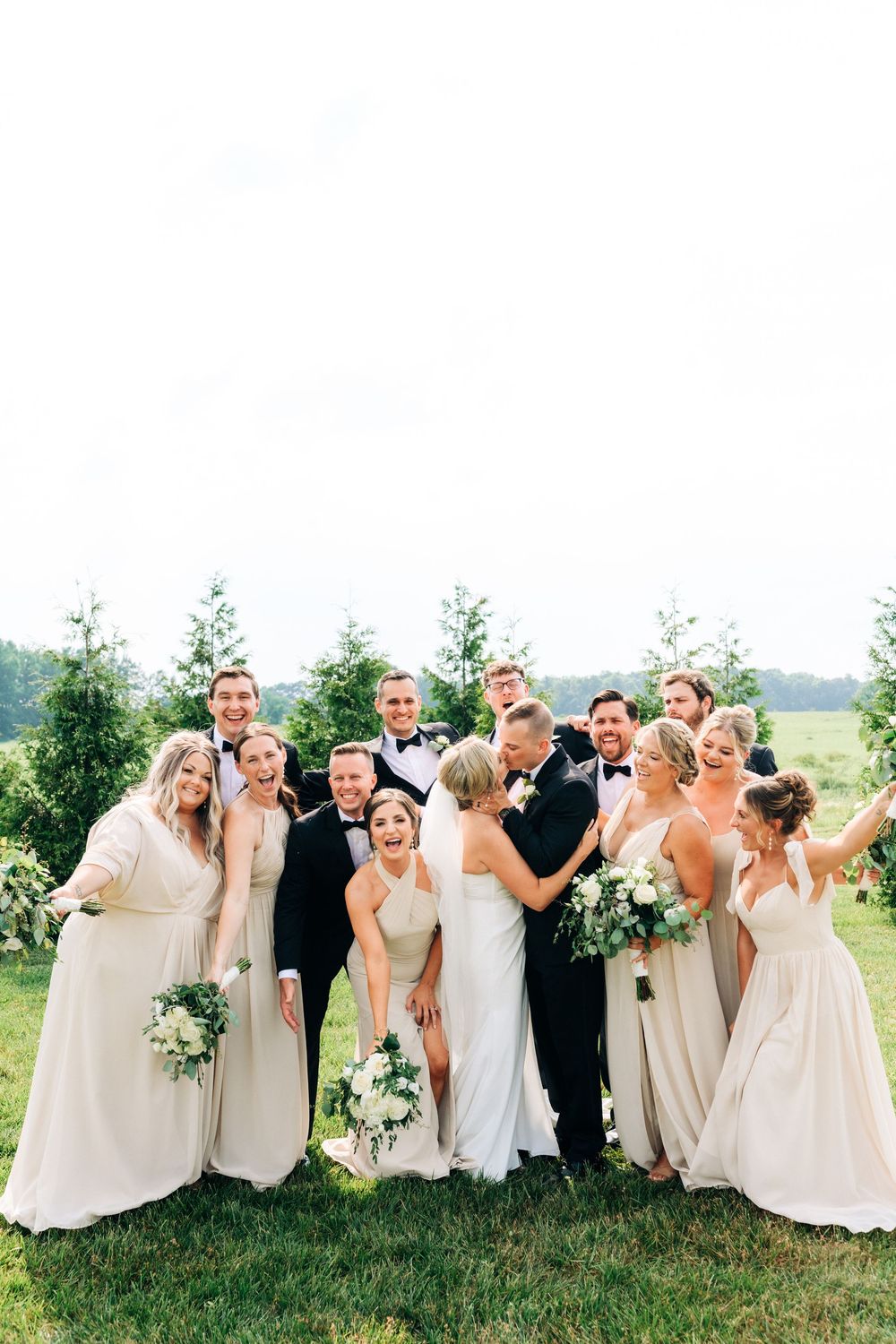 A joyful wedding party poses together outdoors wearing cream and white formal attire.