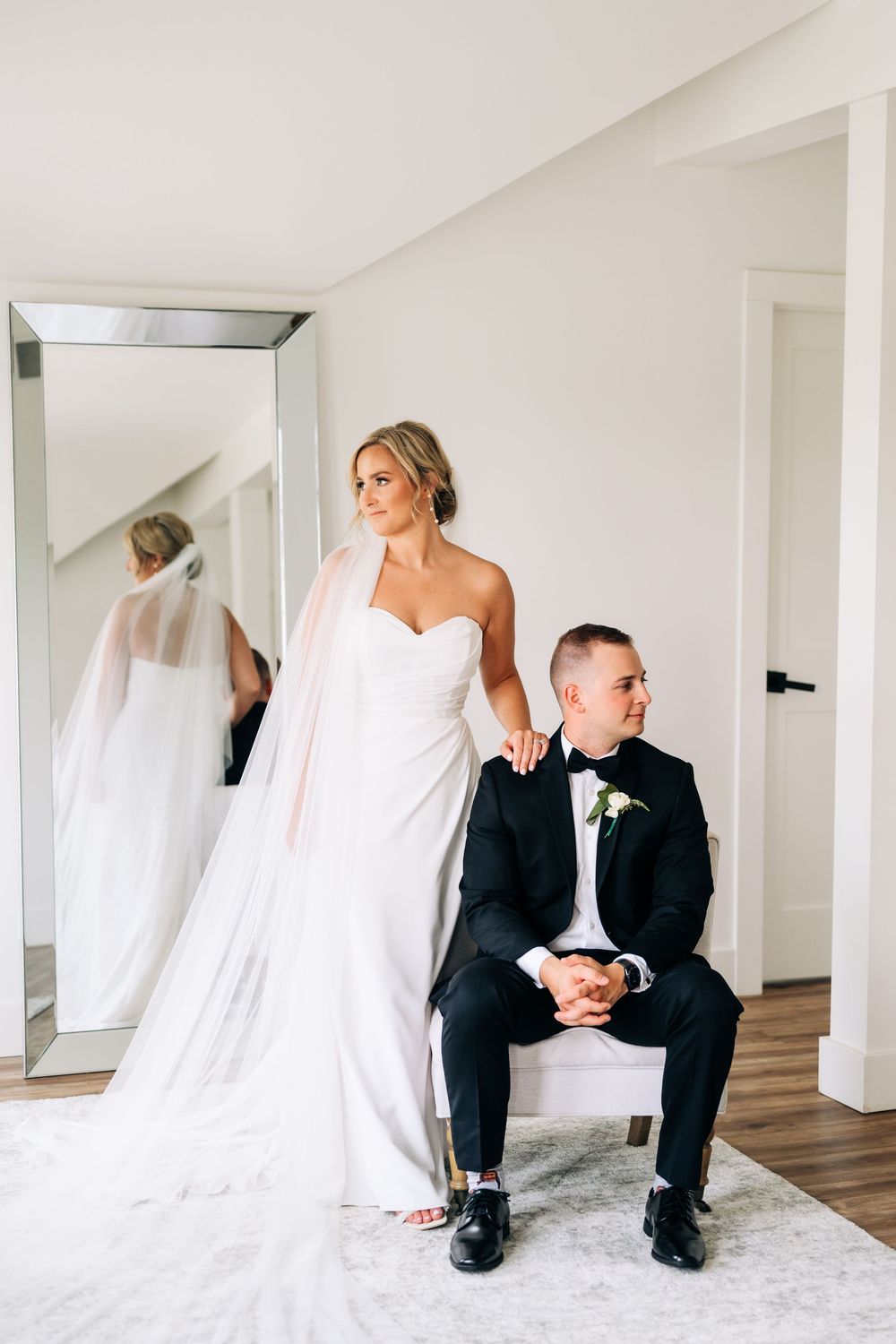Intimate pre-wedding moment with couple in formal attire near a mirror in bright modern room.