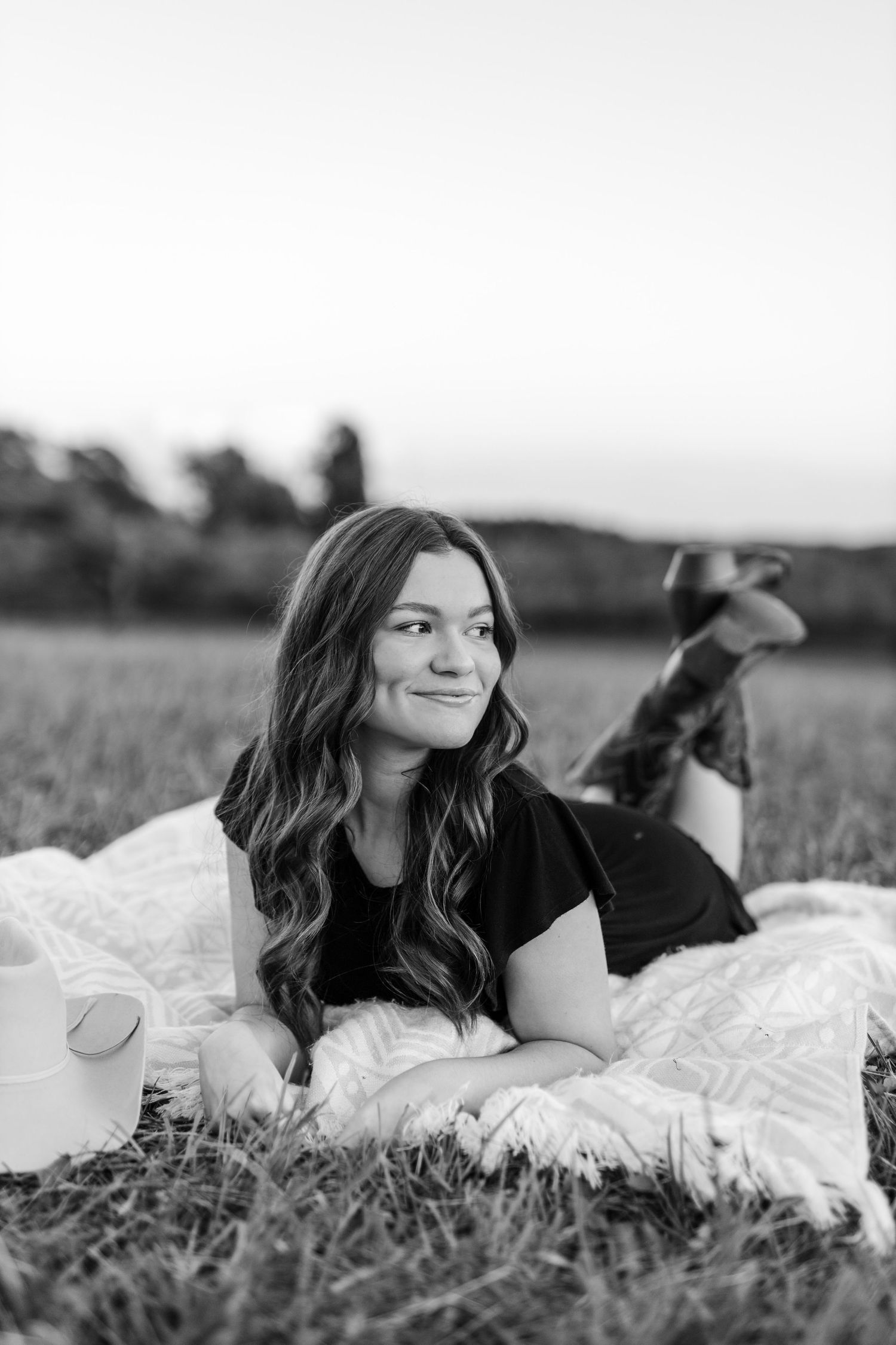 Young woman with wavy brown hair lying on a blanket in a green field during a golden sunset.