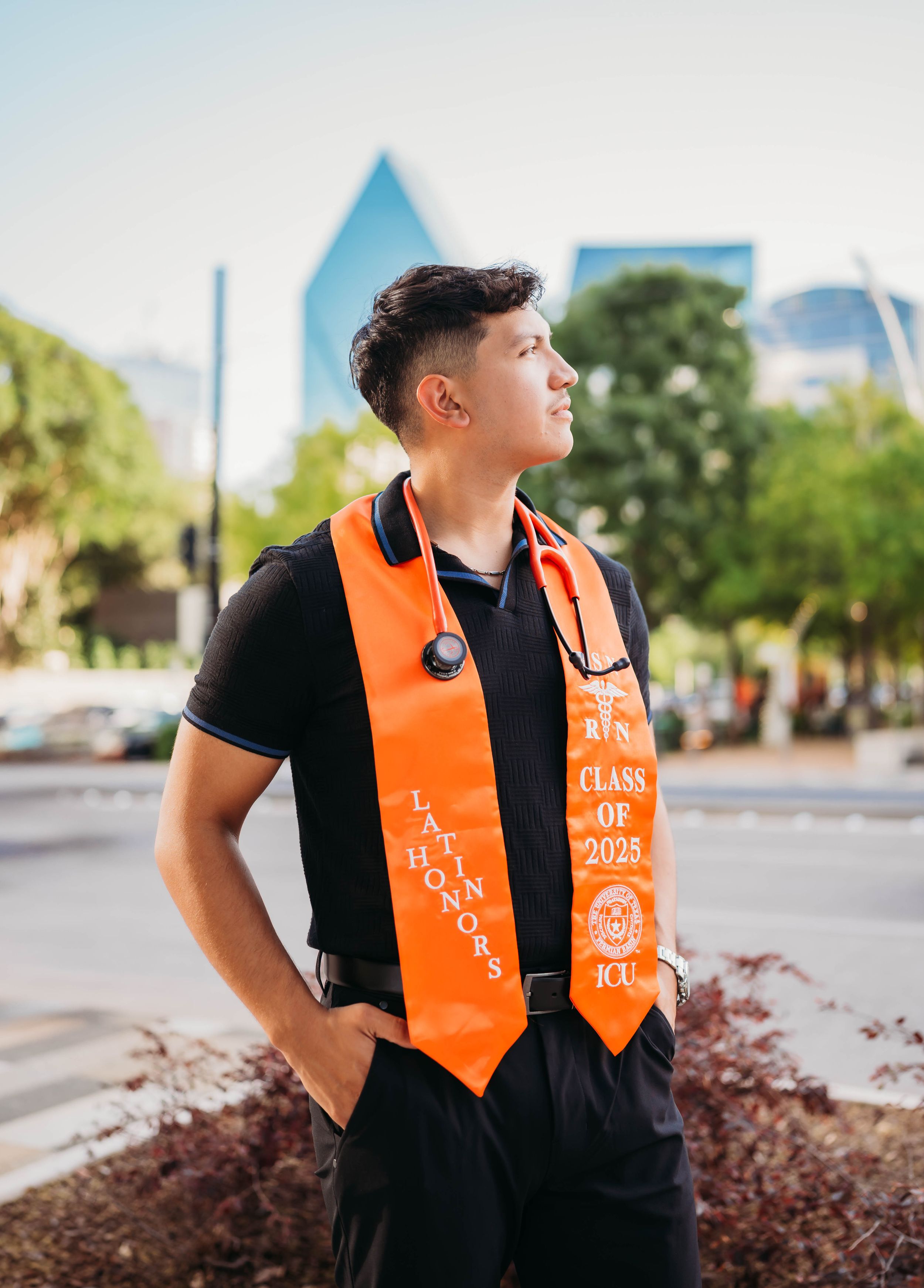 Graduate student wearing black polo and orange graduation stole stands thoughtfully in urban setting with buildings behind them.