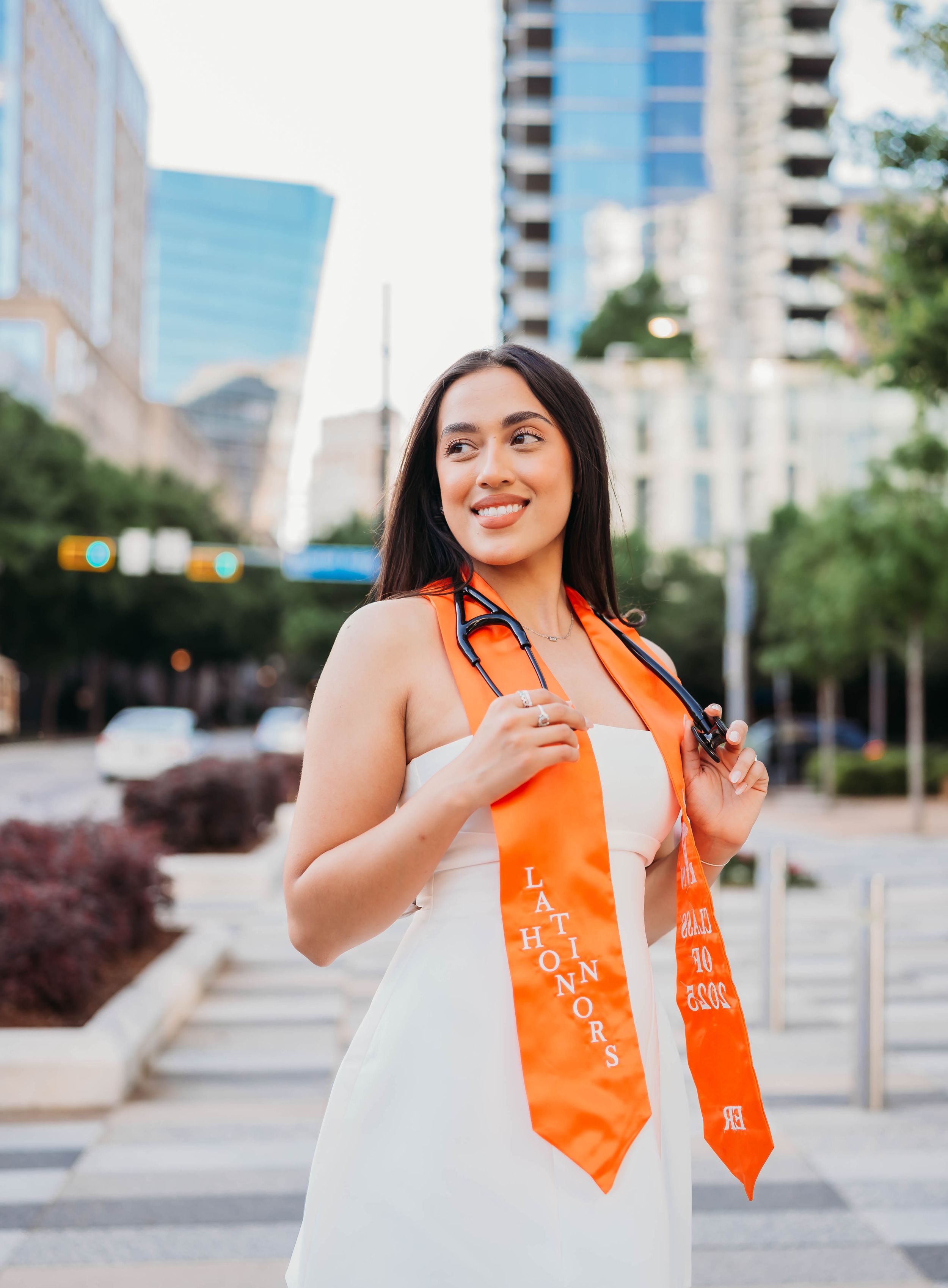 Graduate in white dress with orange sash smiles while standing in urban area with tall buildings in background.