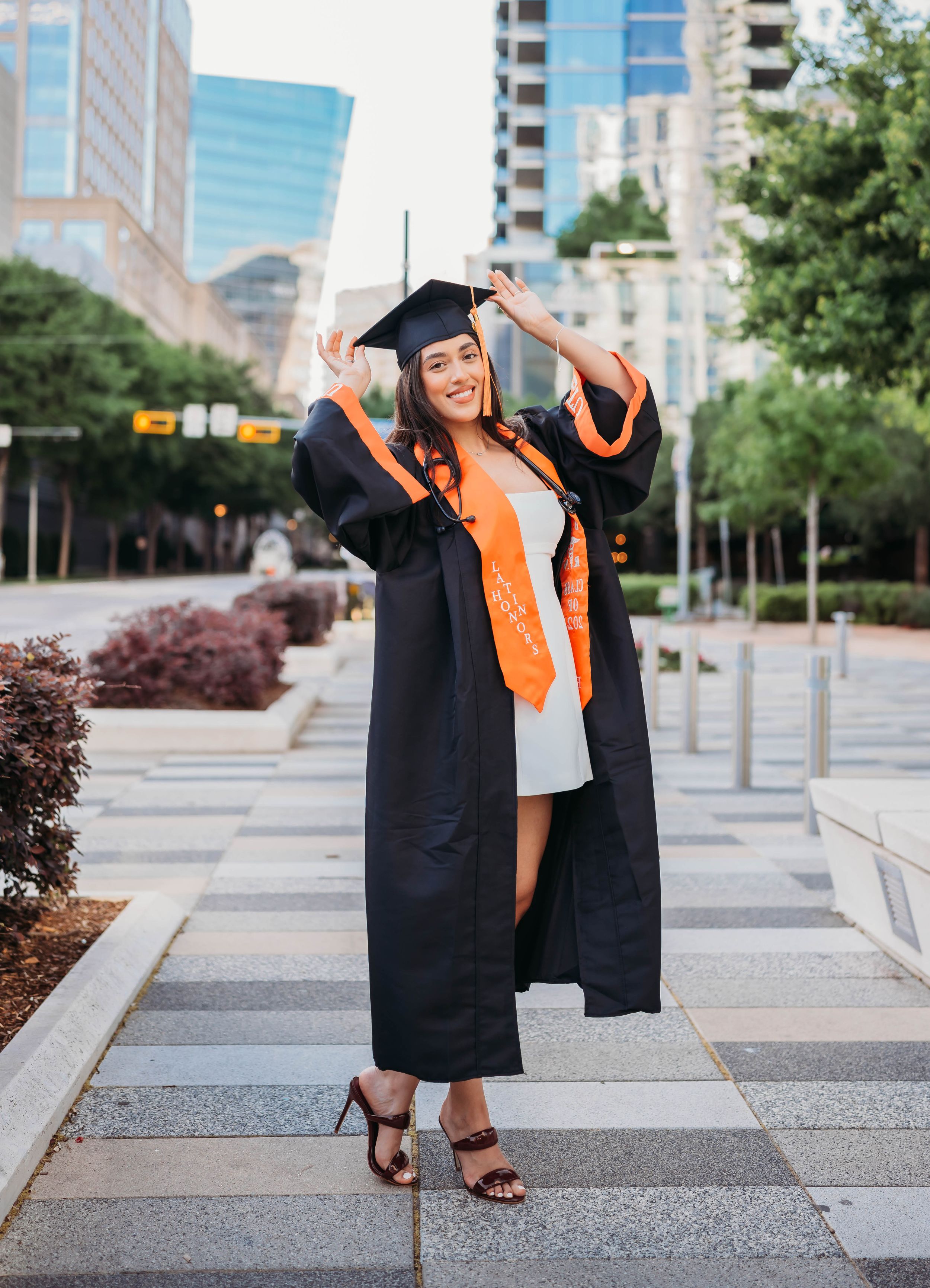 Graduate celebrates with joyful movements in black cap and gown with orange stole on urban plaza.