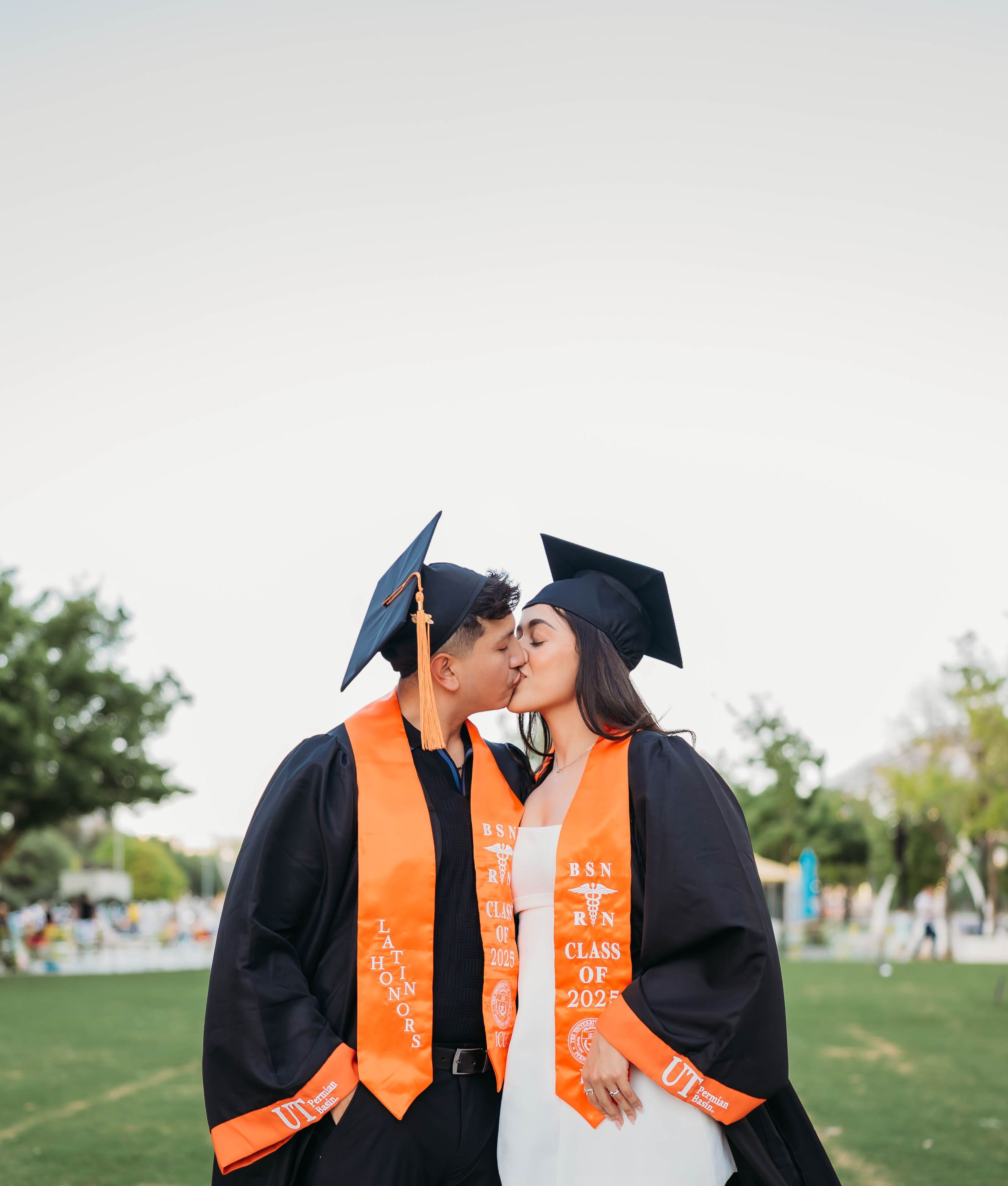 Graduates in black gowns and orange sashes share joyful moments during a graduation photo session outdoors.