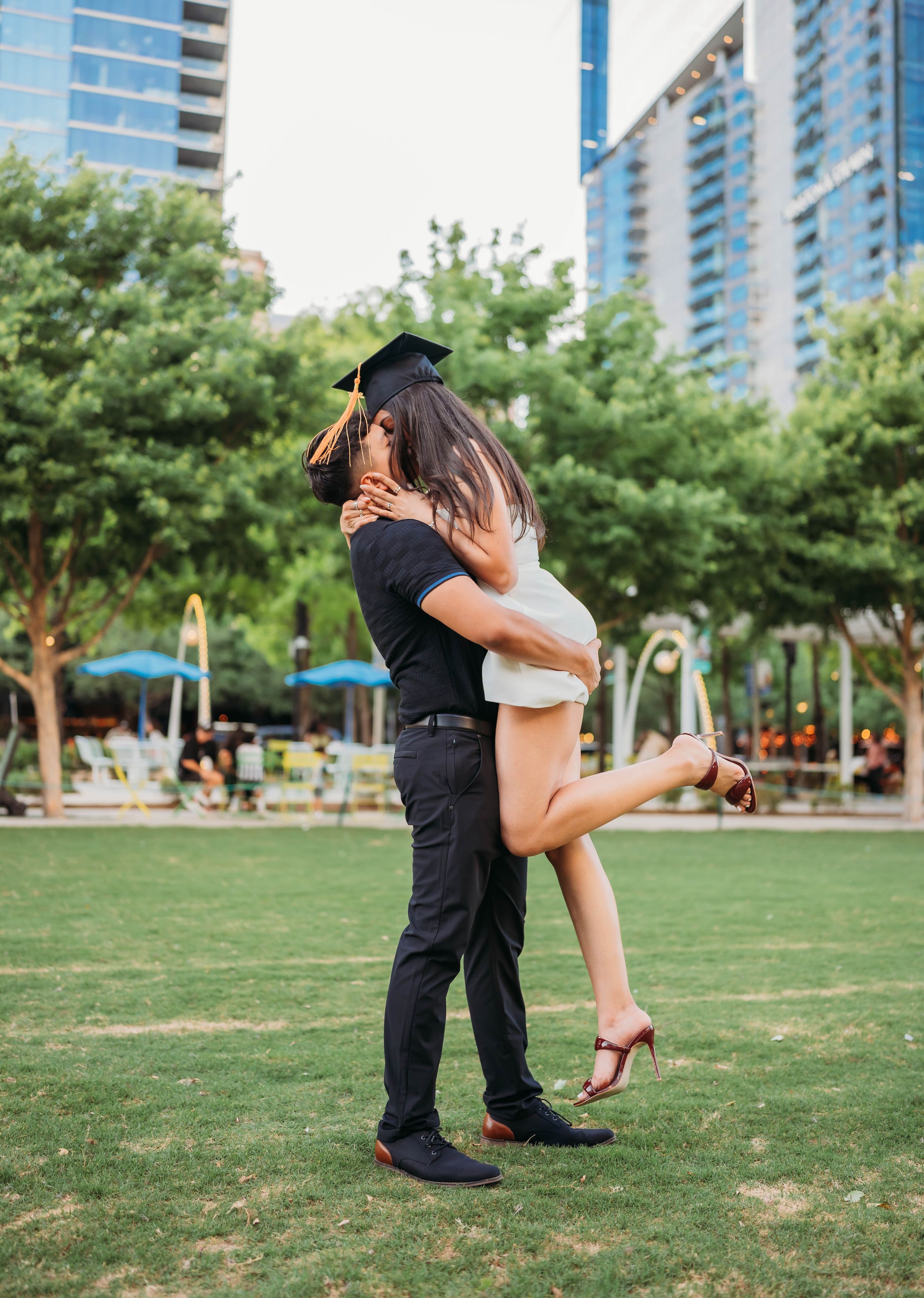 A graduate in cap and gown embraces their partner on a grassy lawn with high-rise buildings in the background.