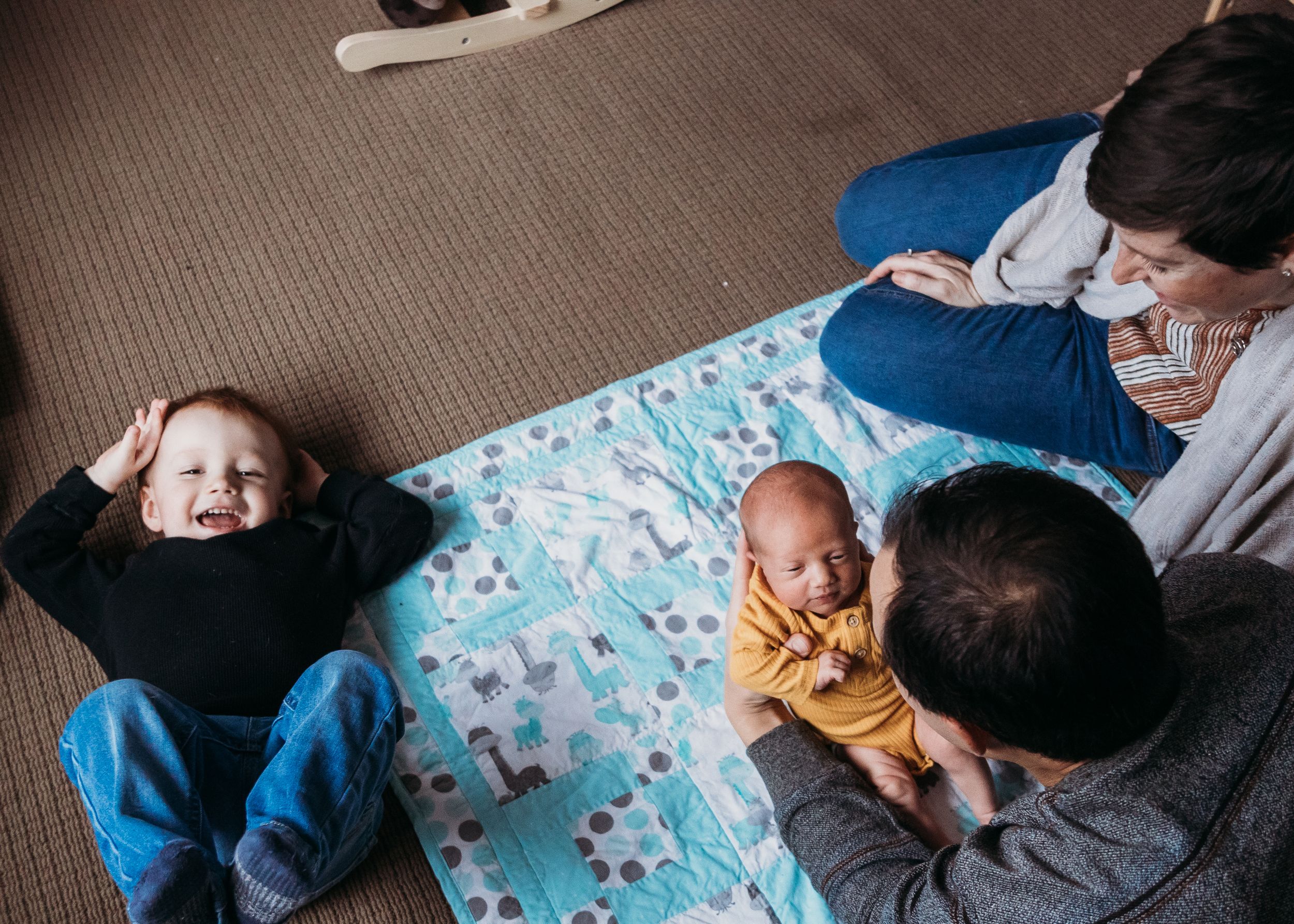 Family lies on patterned blanket while relaxing together on carpeted floor enjoying intimate home time.