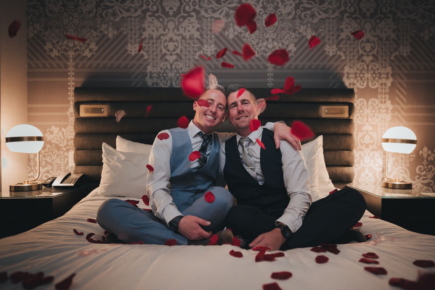 A romantic couple sits on a bed with scattered rose petals on their wedding day.