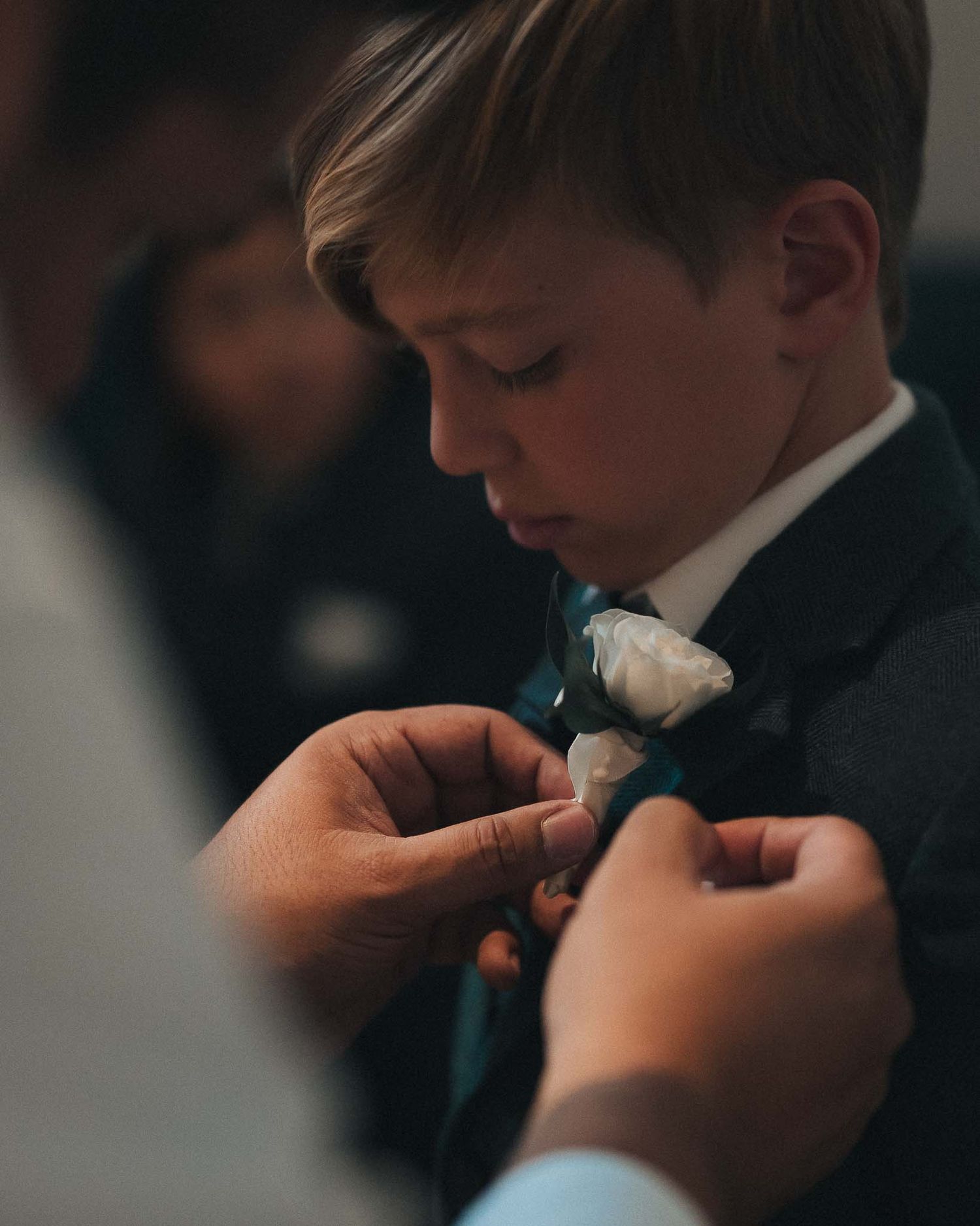 Someone adjusts a white floral boutonniere on a young person wearing a black suit and tie in a formal setting.