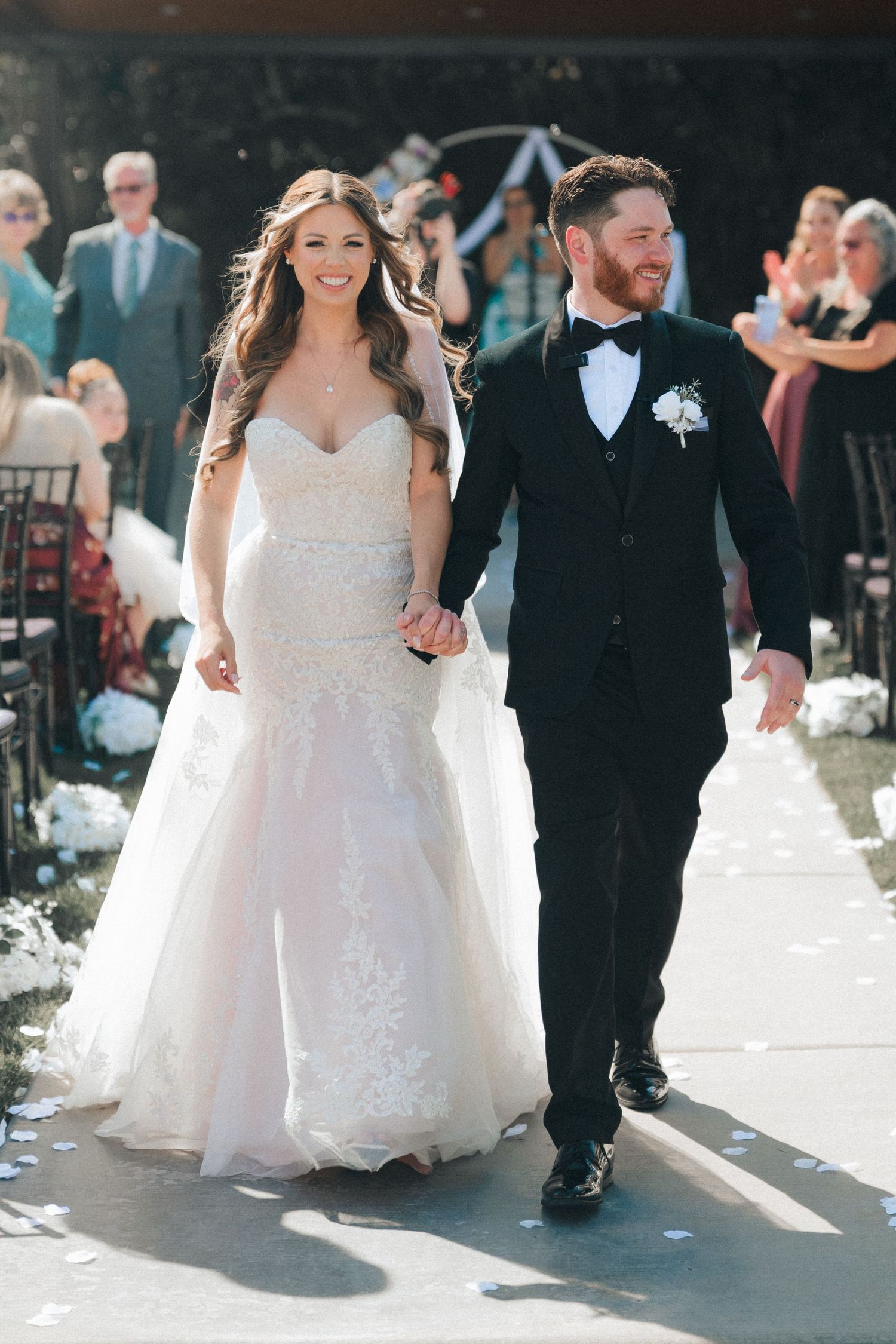 A newlywed couple walks down the aisle on their wedding day, bride in white lace gown and groom in black tuxedo.