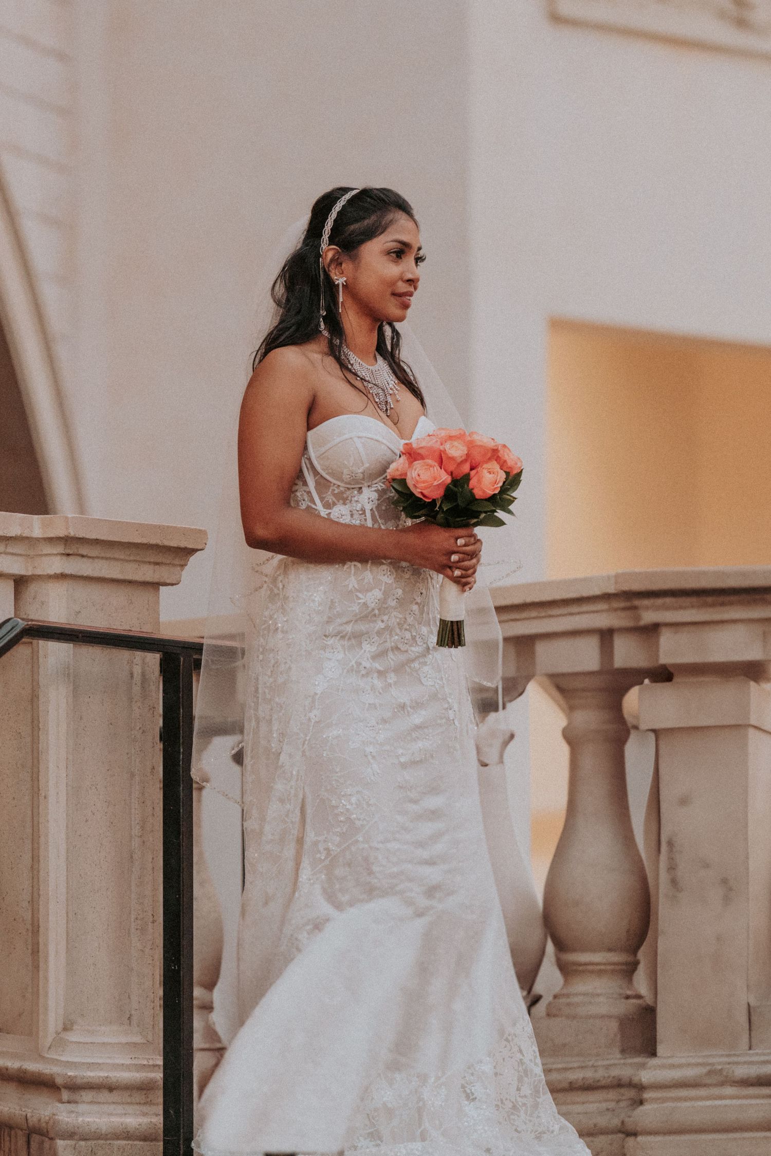 A bride in an elegant white lace wedding gown standing in the Venetian poses against a beige stone colonnade with coral-colored flowers.
