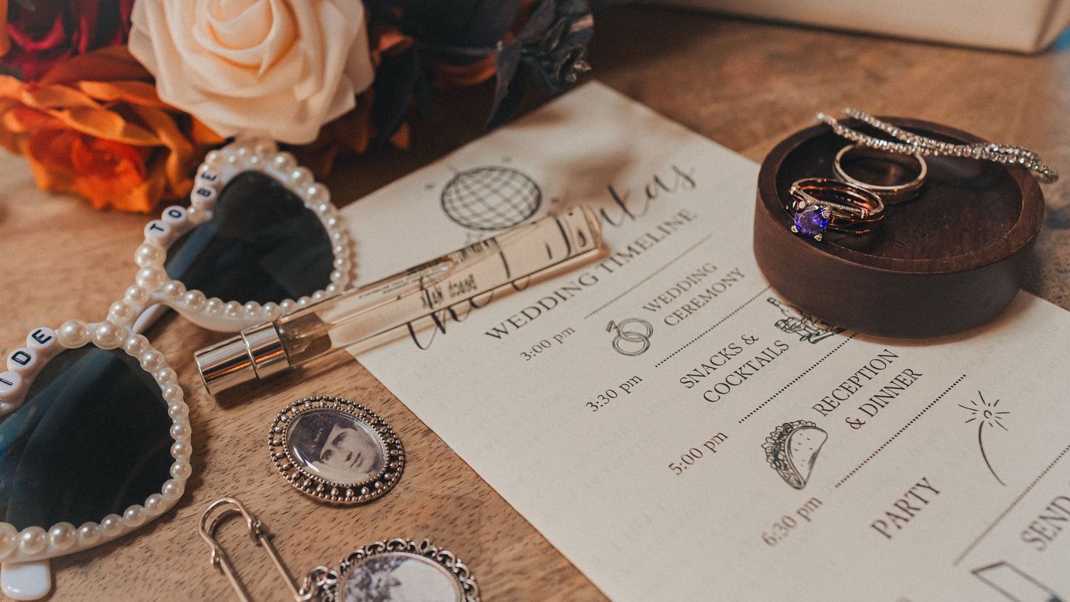 Wedding detail shot with invitation suite with peach roses, gold rings, and vintage-inspired details arranged on wooden surface.