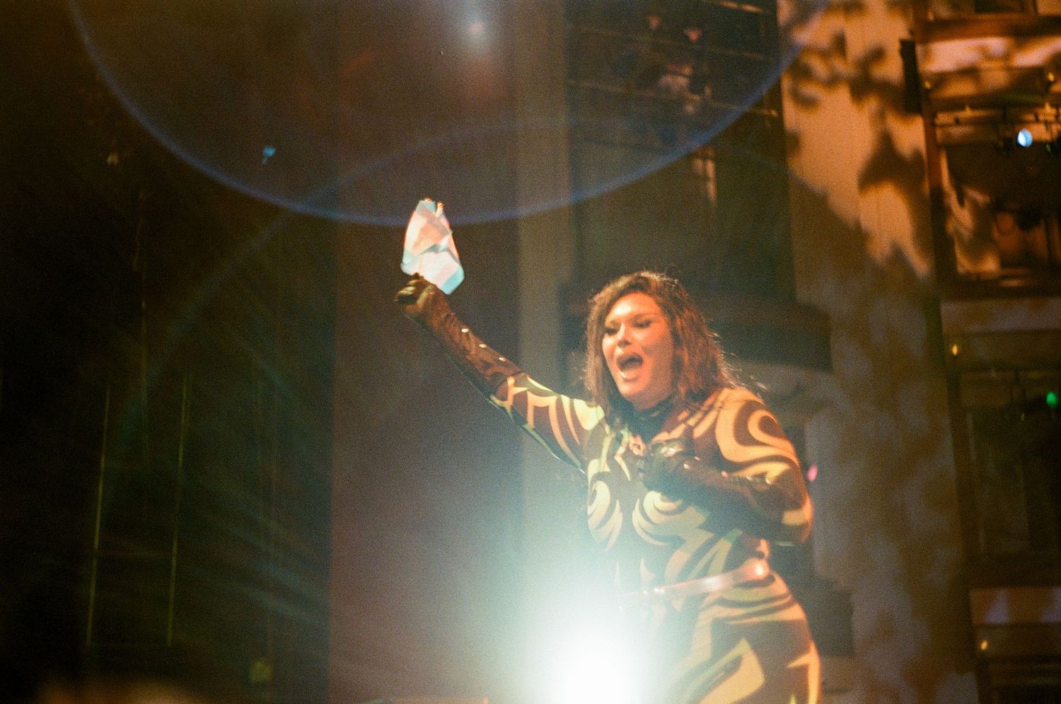 Concert performer in black and gold outfit raises arm triumphantly on stage during dynamic performance with dramatic lighting.
