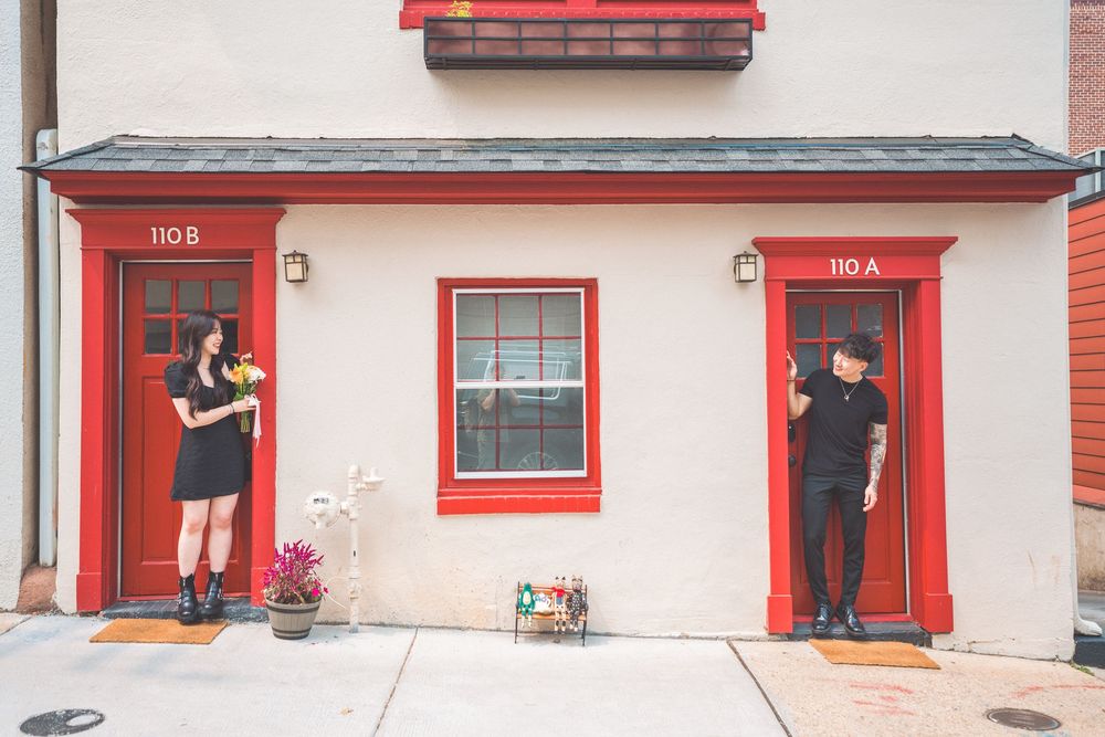 Couple stands at separate red doors of white building for creative photo.