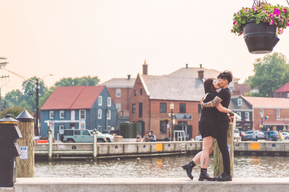 Couple embraces on waterfront with historic buildings and harbor in background.