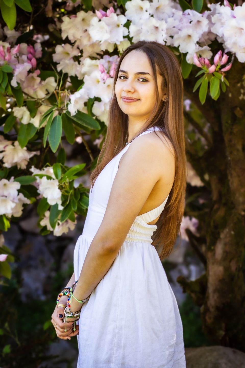 A person in a white summer dress poses outdoors against blooming white and pink rhododendron flowers.