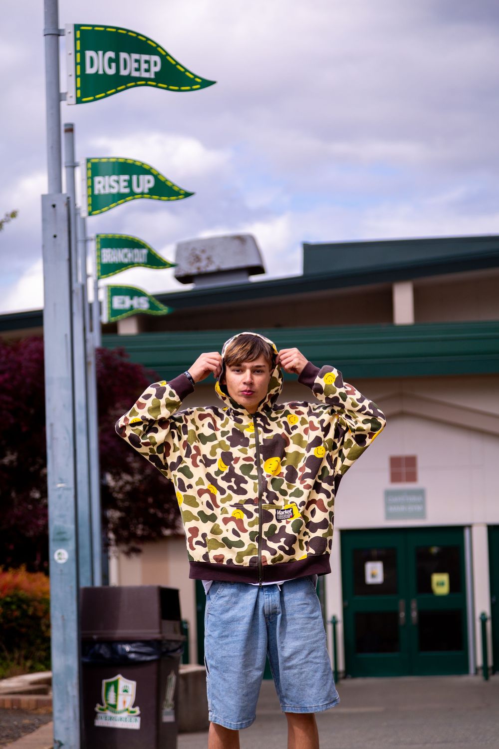 Person in camo patterned hoodie and denim shorts stands near green flags at a golf facility entrance.
