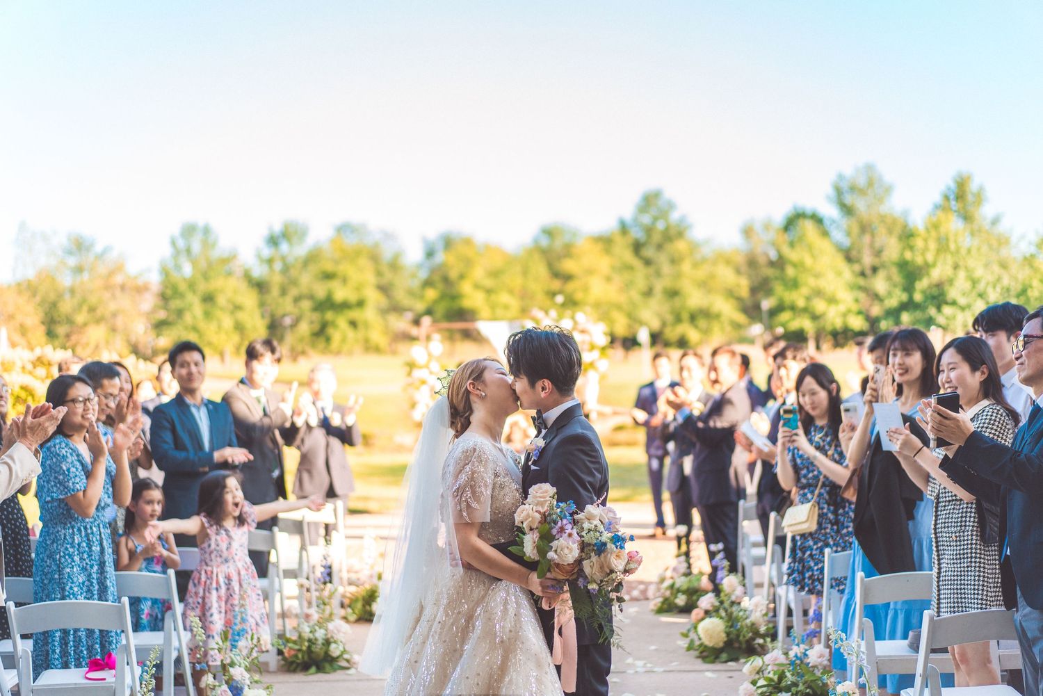 Wedding guests applaud during an outdoor ceremony with blue chairs and autumn trees in the background.
