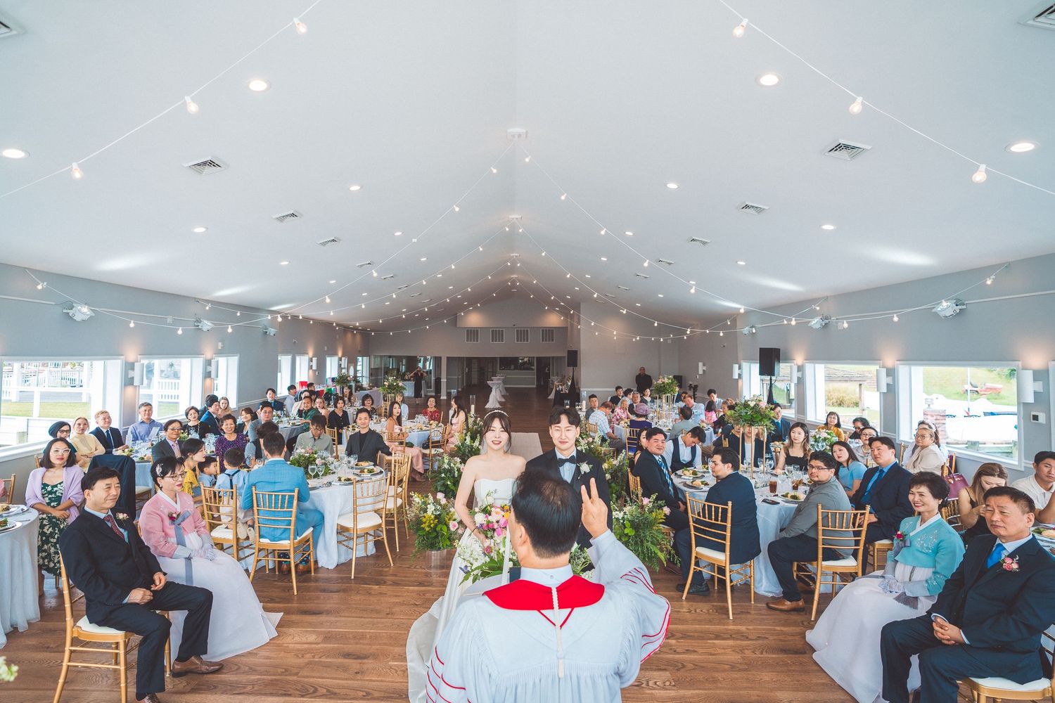 Wide view of wedding guests seated in a spacious venue with high ceilings and elegant lighting.