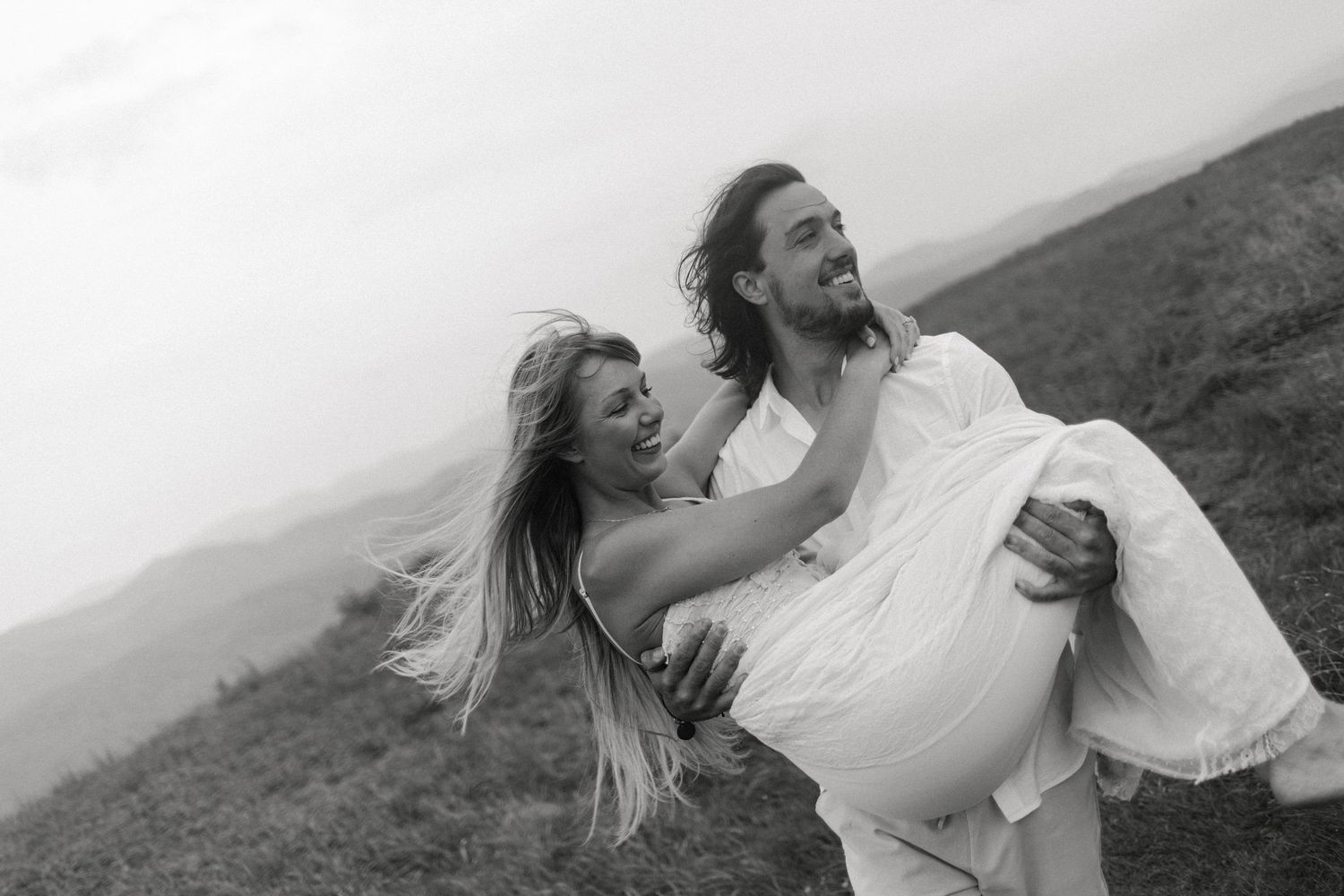A joyful couple embraces while twirling on a hillside in black and white photography.