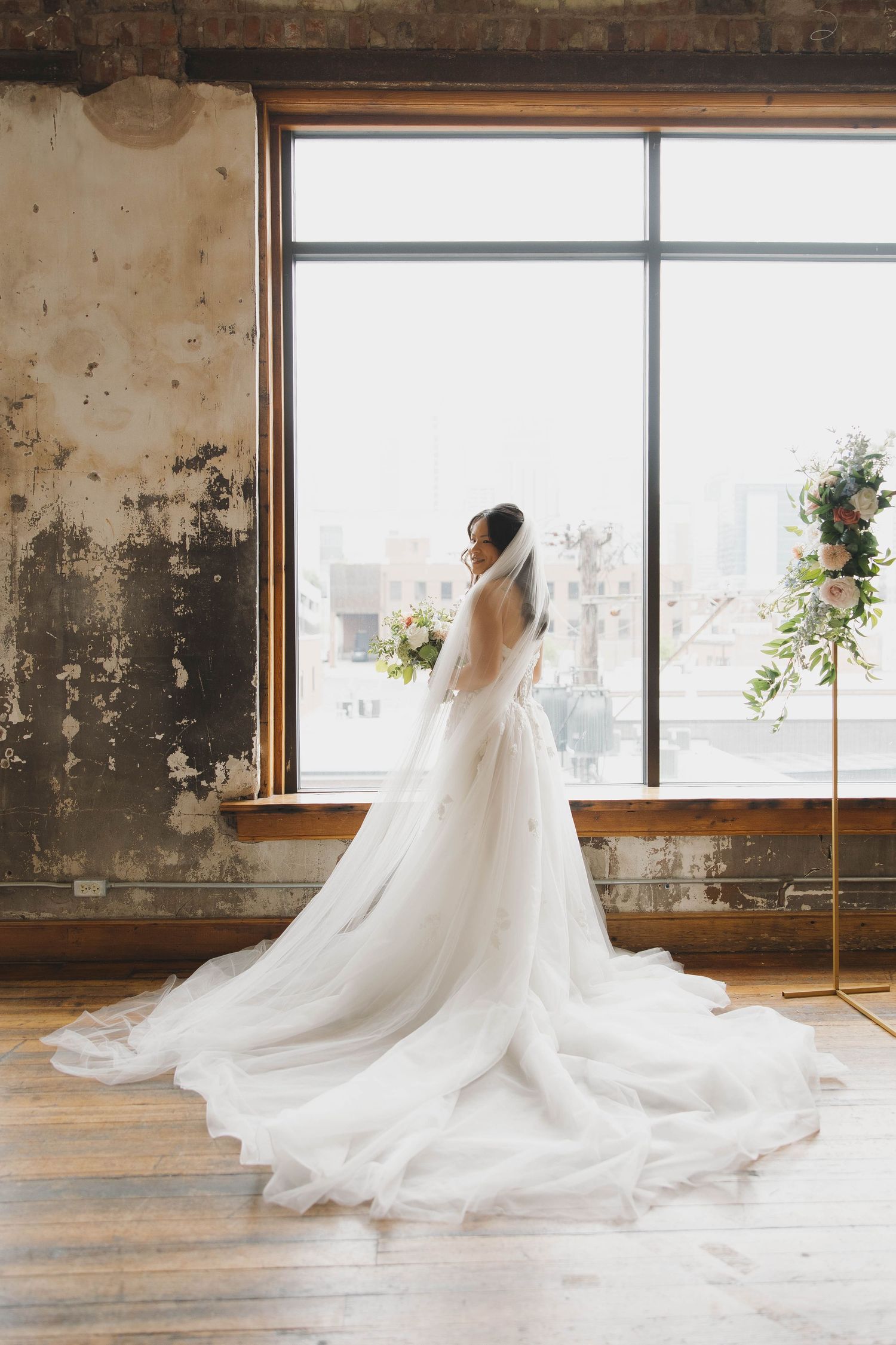 A bride in an elegant white wedding gown and veil holds a delicate bouquet while standing by a bright window.