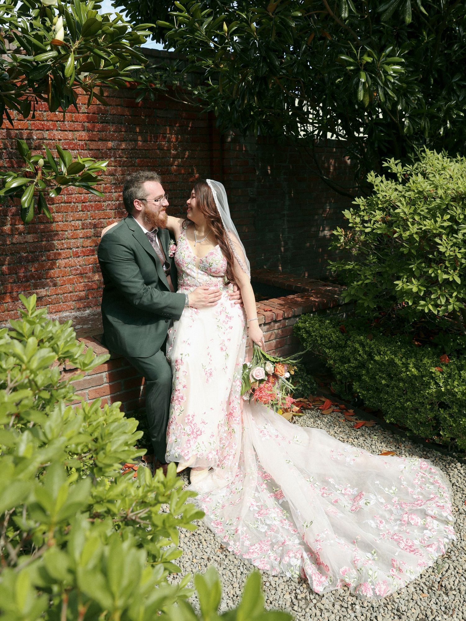 A couple in wedding attire embrace in a garden setting with brick walls and lush greenery.