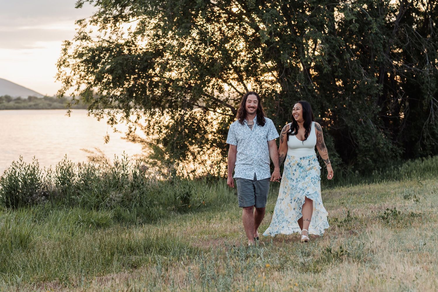 Engaged couple walks along Columbia River at Chiawana Park during sunset as golden light filters through trees in Pasco, Washington.